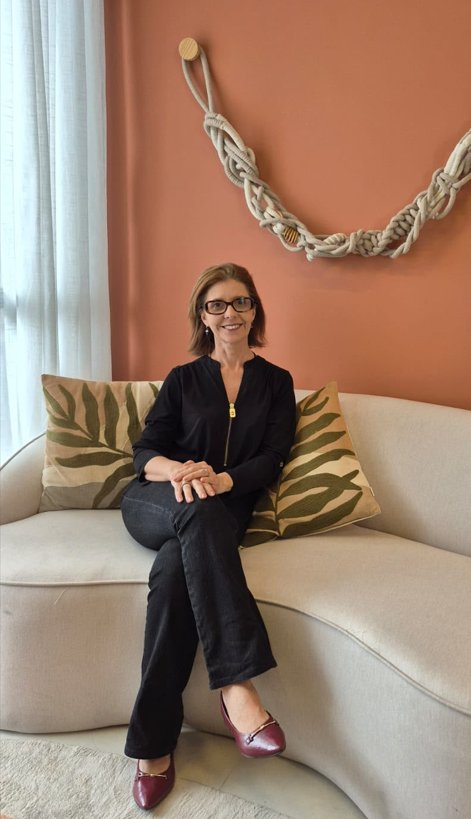 Fernanda Familiar sentada em um sofá claro de linhas curvas, com almofadas de folhagem e parede terracota.